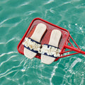 A pair of cream-colored slide sandals with navy blue ruffle straps and white floral trim, resting on a red scoop in turquoise water.
